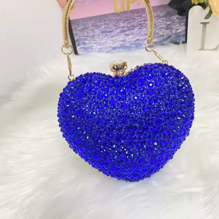 Bolsa clutch festa azul em formato de coração, cravejada com cristais azuis vibrantes, alça metálica dourada estruturada e fecho superior com detalhe em pedra – loja Bolsa Clutch