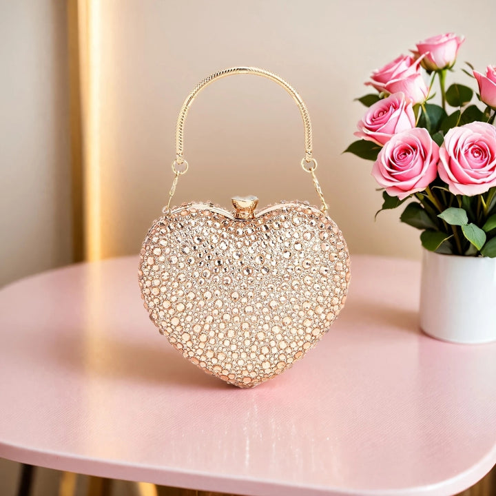 Em cima de uma mesa ao lado de flores está a Bolsa de festa clutch Feminina Champagne em formato de coração, cravejada com cristais azuis vibrantes, alça metálica dourada estruturada e fecho superior com detalhe em pedra – loja Bolsa Clutch