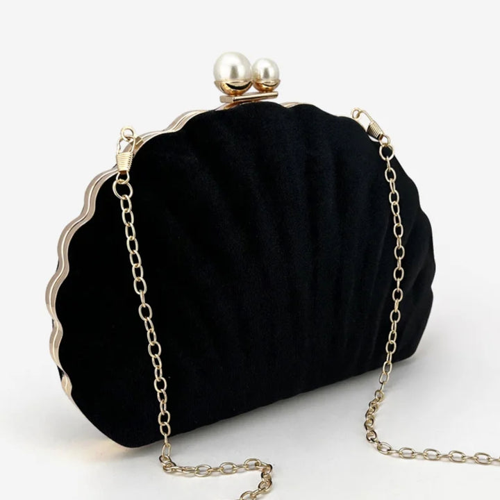Vista diagonal da Bolsa clutch festa preta concha luxuosa, em veludo, com formato de concha e bordas douradas onduladas. Fecho superior com duas pérolas e alça de corrente fina dourada. Ideal para casamentos, formaturas e eventos elegantes.