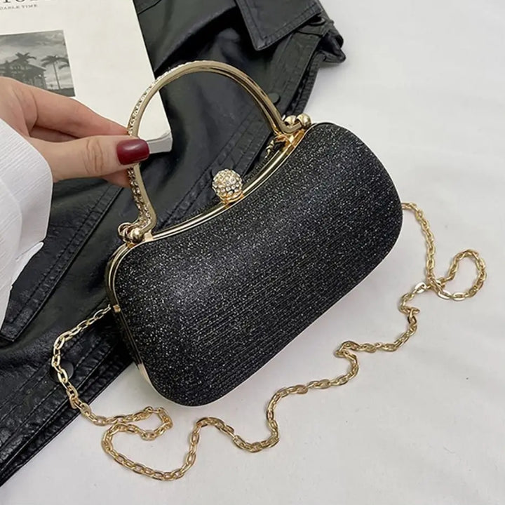Bolsa clutch festa preta elegance com brilho sutil, design retangular arredondado. Alça de mão superior dourada com detalhes em strass, fecho de esfera cravejada e alça de corrente fina dourada. Ideal para festas e eventos formais. Fundo de mesa branca com uma jaqueta de couro ao lado. Loja Bolsa Clutch
