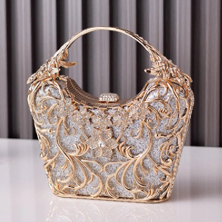 Bolsa clutch de festa prata com acabamento metálico dourado rendado, alça rígida escultural e aplicação de flores em strass, design exuberante e sofisticado – loja Bolsa Clutch