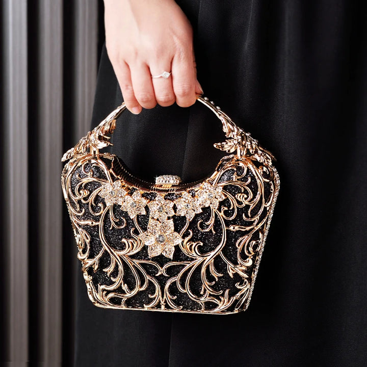 Mulher segurando a Bolsa clutch festa preta com acabamento metálico dourado rendado, alça rígida escultural e aplicação de flores em strass, design exuberante e sofisticado – loja Bolsa Clutch