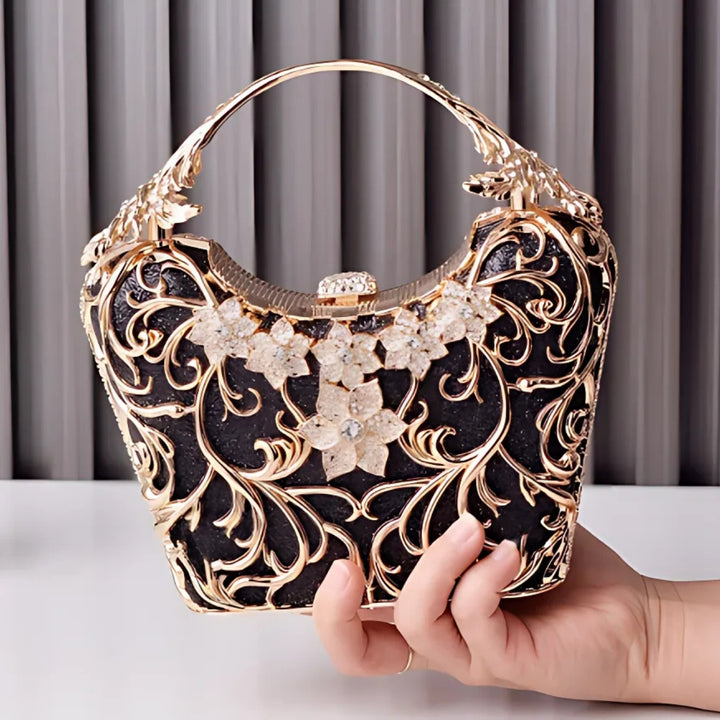 Mão feminina segurando a Bolsa clutch festa preta com acabamento metálico dourado rendado, alça rígida escultural e aplicação de flores em strass, design exuberante e sofisticado – loja Bolsa Clutch