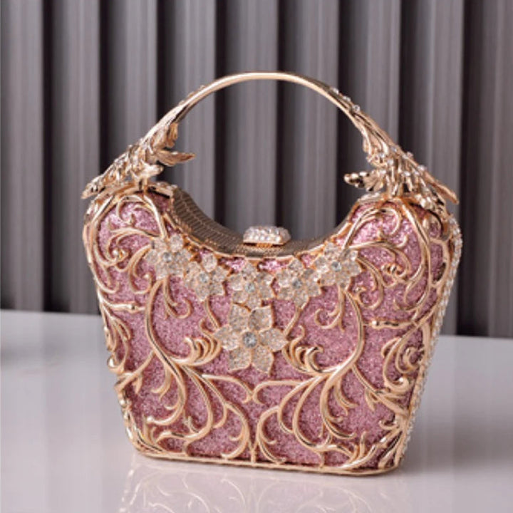 Bolsa de Festa clutch na cor rosa com detalhes dourados com acabamento metálico rendado, alça rígida escultural e aplicação de flores em strass, design exuberante e sofisticado – loja Bolsa Clutch