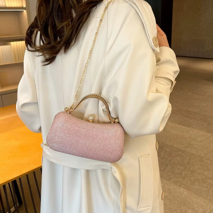 Mulher usando com alça de ombro a Bolsa feminina de mão clutch de festa rosa elegance com brilho, design retangular arredondado. Alça de mão superior dourada com strass, fecho de esfera cravejada e alça de corrente fina. Perfeita para eventos especiais e festas.