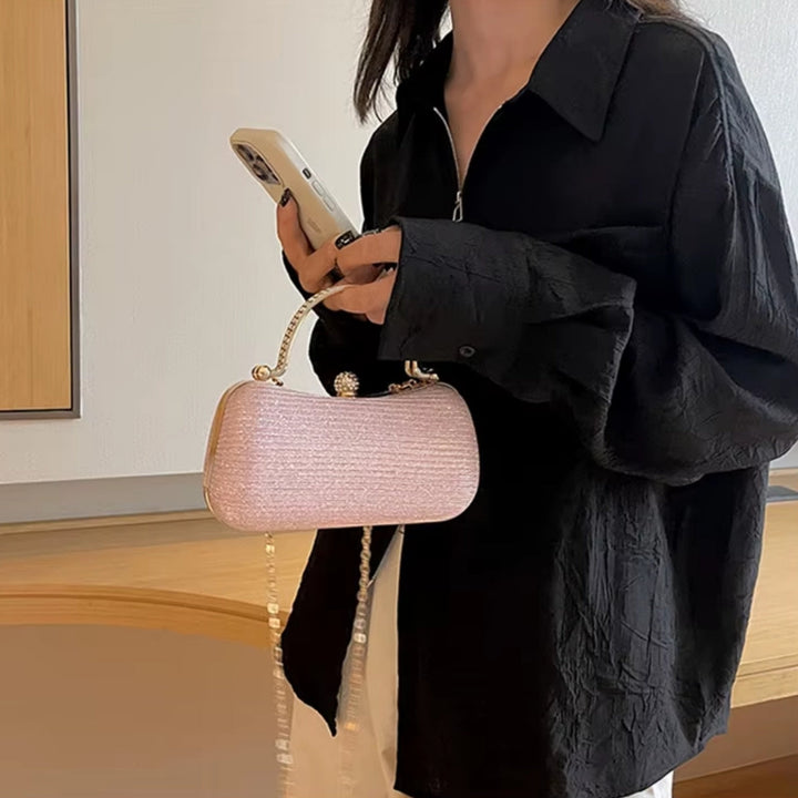 Mulher com a Bolsa clutch festa rosé gold elegance com brilho, design retangular arredondado. Alça de mão superior dourada com strass, fecho de esfera cravejada e alça de corrente fina. Perfeita para eventos especiais e festas.
