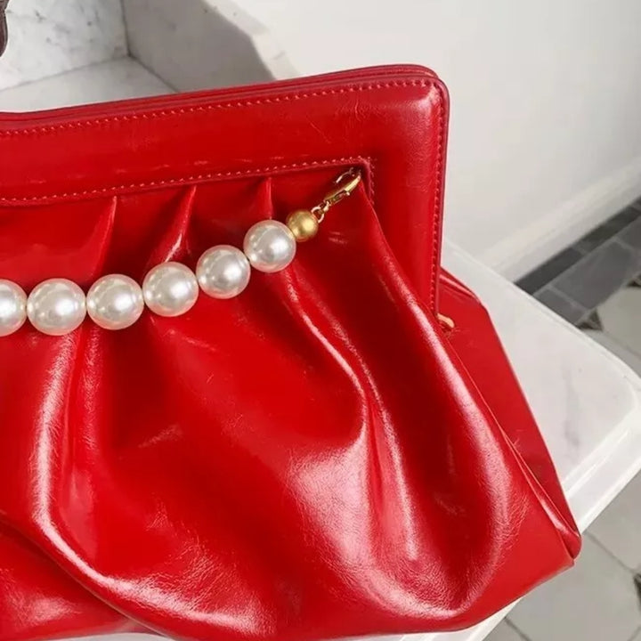 Detalhe lateral da alça perolada da Bolsa clutch festa vermelha vibrante, em material sintético com efeito drapeado e alça superior fixa de pérolas brancas com terminais dourados. Possui fechamento seguro. Ideal para adicionar um toque de cor e glamour a eventos especiais.