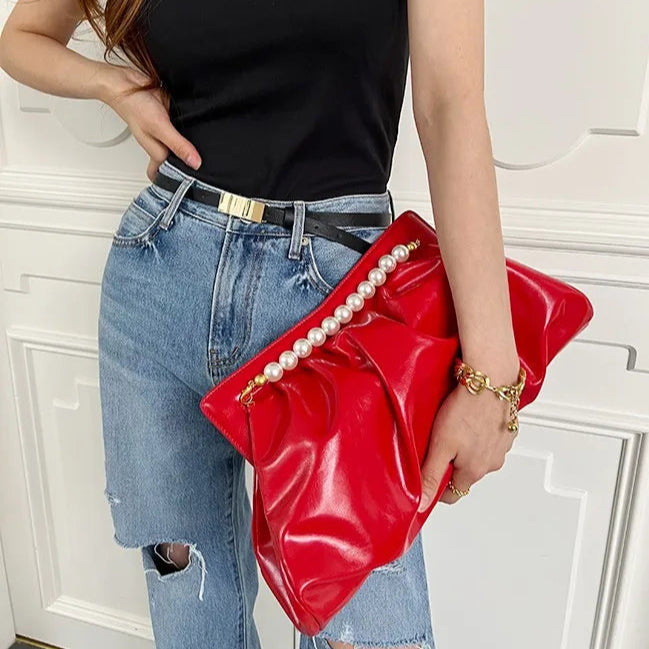 Mulher segurando a Bolsa clutch festa vermelha vibrante, em material sintético com efeito drapeado e alça superior fixa de pérolas brancas com terminais dourados. Possui fechamento seguro. Ideal para adicionar um toque de cor e glamour a eventos especiais.