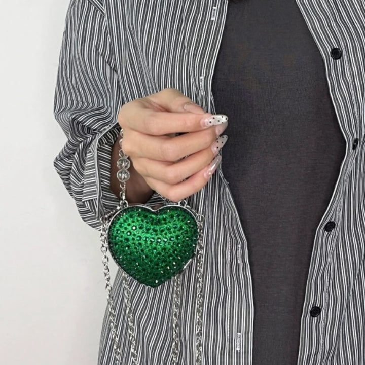 Mão feminina segurando a Bolsa clutch festa Verde em formato de coração, cravejada com cristais azuis vibrantes, alça metálica dourada estruturada e fecho superior com detalhe em pedra – loja Bolsa Clutch